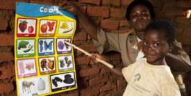 This is a child at Lonjezo CBCC in Dowa district, central region of Malawi pointing at a color matching poster held against a brick wall by a female educator